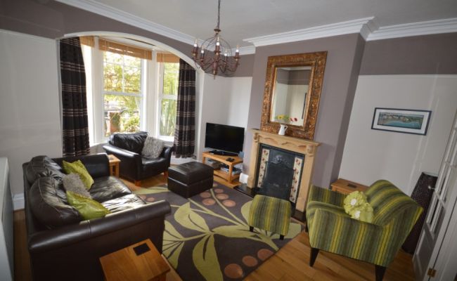 Image of the living room. Large space with two arm chairs and one long sofa. Has a large TV in the corner of the room